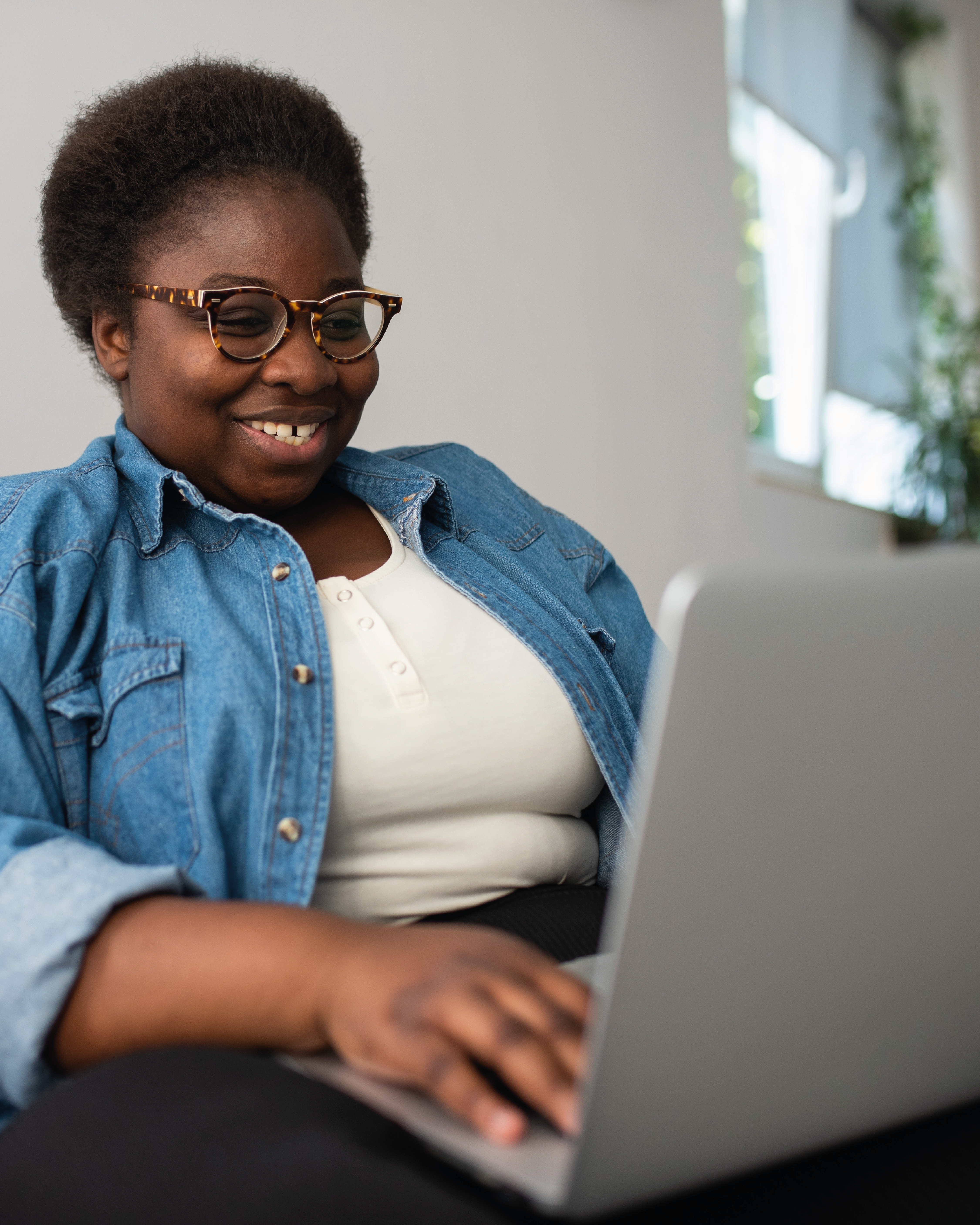 A Black person with short hair working on their laptop; they're wearing a white t-shirt, a denim shirt and glasses