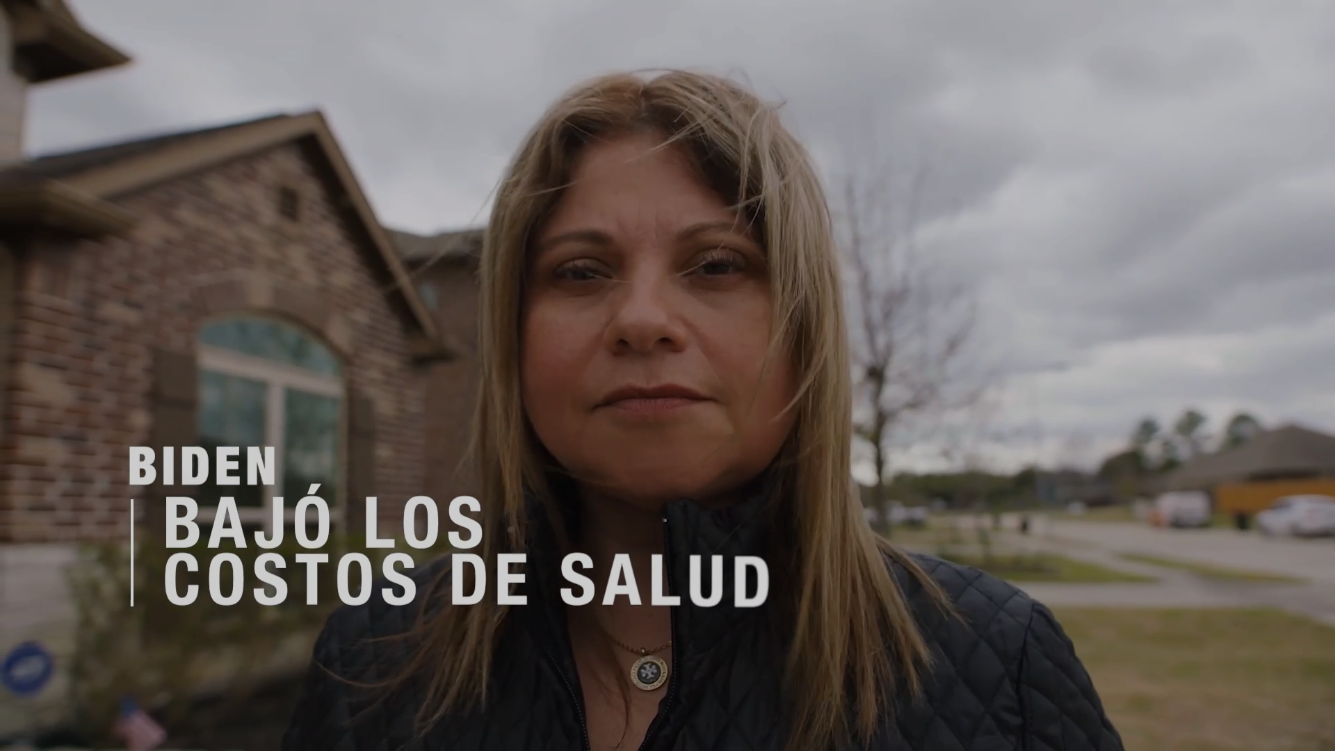 A medium close-up portrait of a woman looking at camera standing outside of a house, and text on screen that reads "Biden bajó los costos de salud"