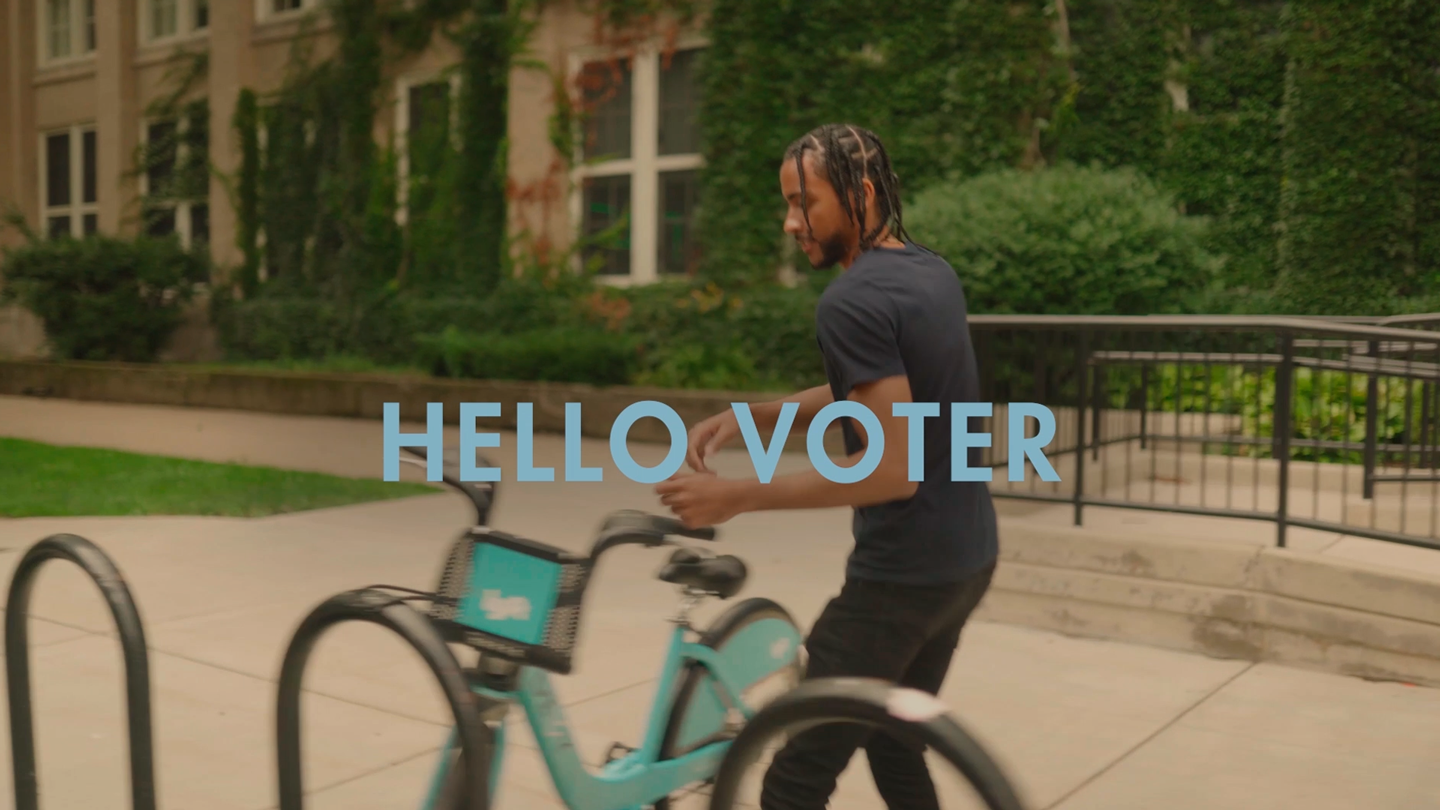 A Black man who is about to get on a bike outside of a building with a lot of plants. Text on screen reads "HELLO VOTER"